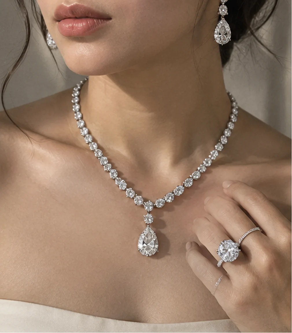Diamond rivière necklace with a pear-shaped drop, diamond drop earrings, and a hand at the collarbone wearing an oval solitaire