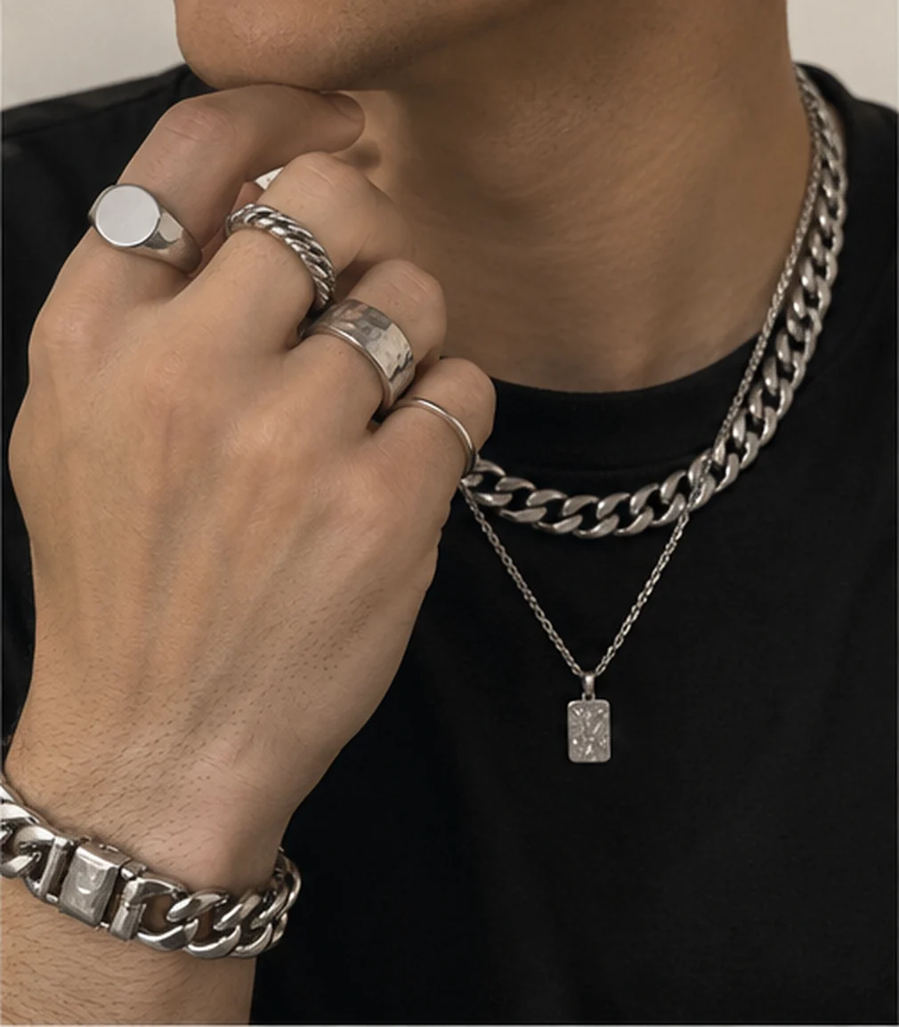 Man in a black tee, hand raised at his jaw wearing a silver signet, rope, hammered and stacking rings, a heavy curb bracelet and layered silver chains