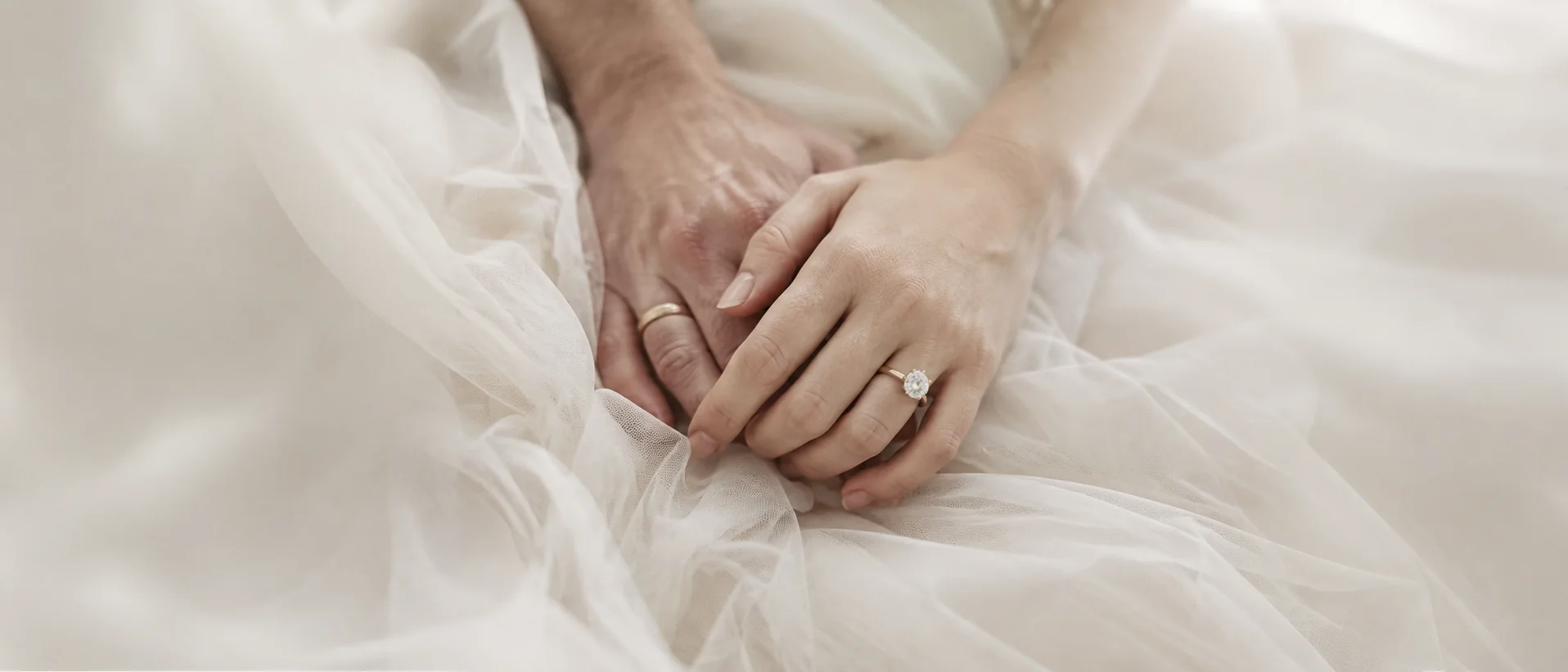 Two generations of hands resting together on ivory tulle — a gold wedding band and a diamond solitaire